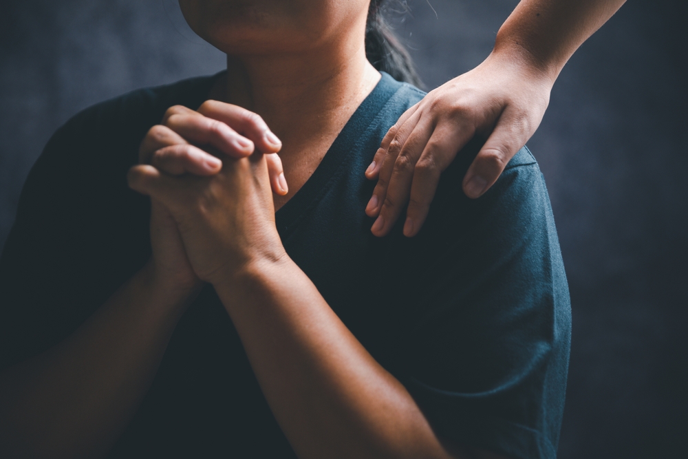 A woman prays while another rests a hand on her shoulder.