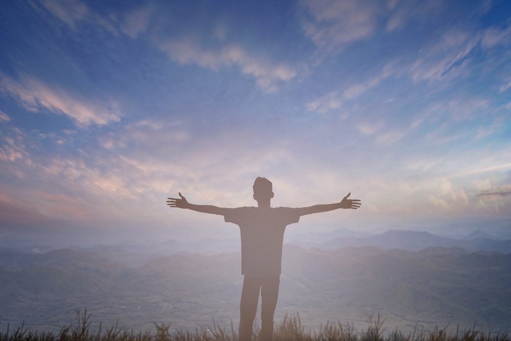 A man stands on a hazy hillside with arms outstretched.