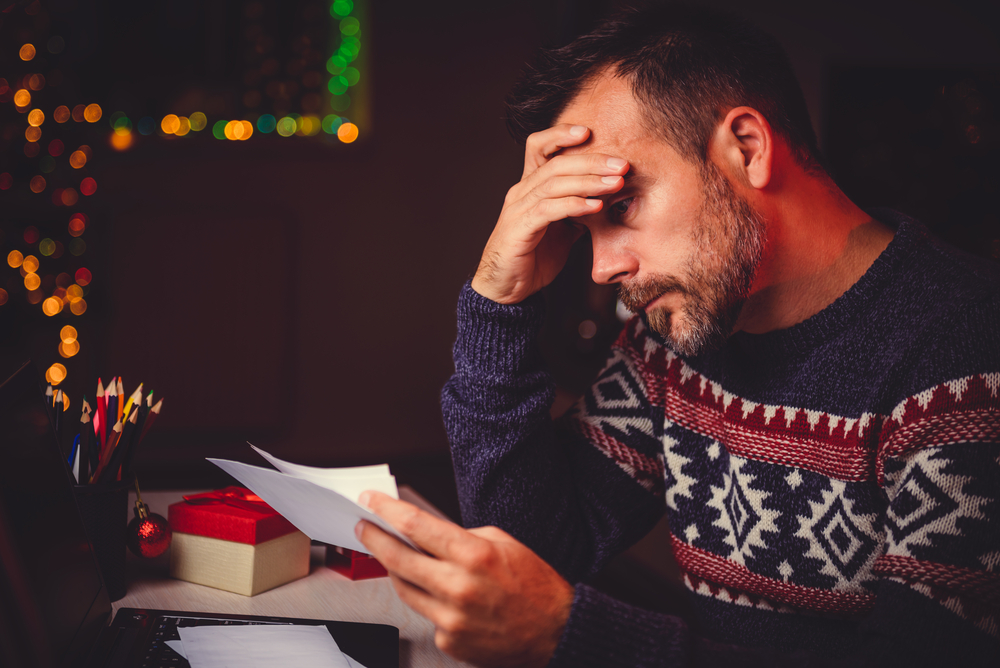 A man in a Christmas sweater reads a letter with an unhappy expression.