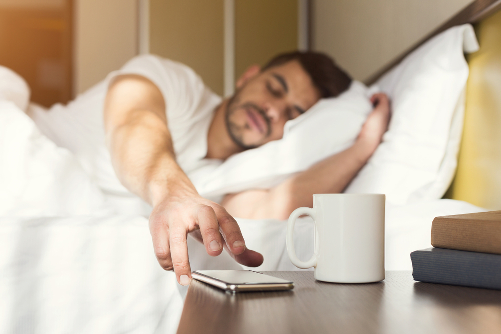 A man reaches for his phone on his bedside stand. 