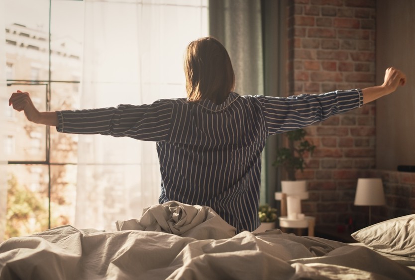 A woman stretches her arms on the side of her bed.